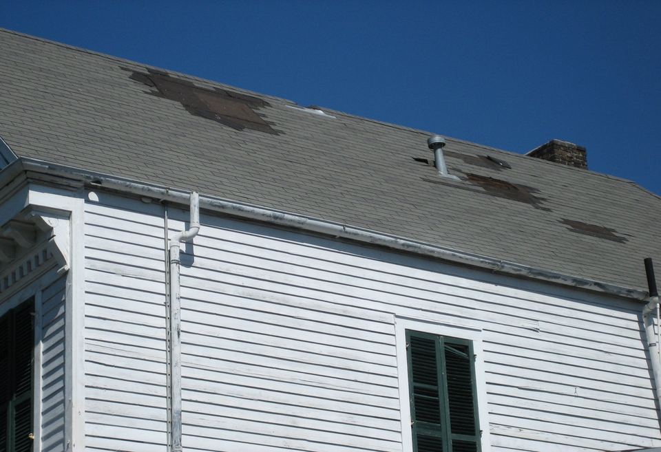 White clapboard house with patches of shingles torn off its gray roof by wind