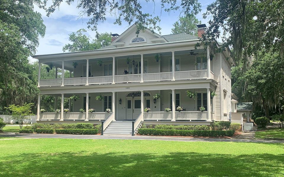 White two-story house with double front porches, a low hipped roof and moss-draped oaks