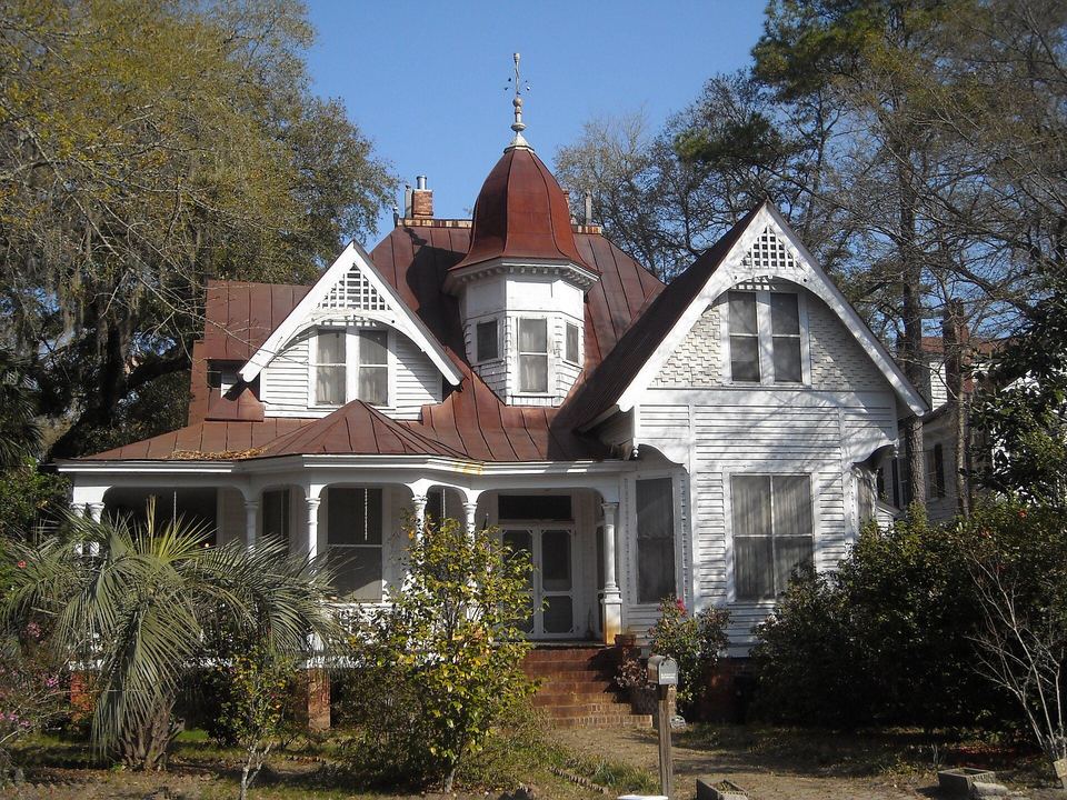 White Victorian house with a rust-red metal roof, turret and wraparound porch among palmettos