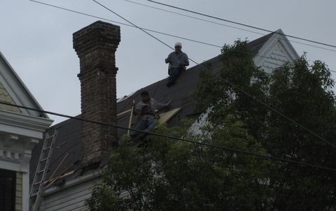 Two roofers working on a steep dark roof beside a tall brick chimney