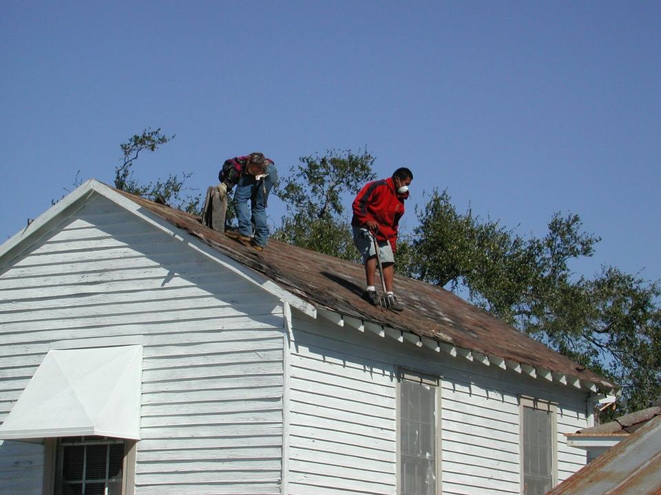 Two roofers stripping old shingles from a white clapboard house beneath a live oak